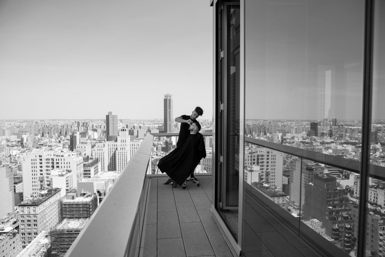 Julien Howard a.k.a. Velo Barber cutting a person's hair high up on the deck of a sky scraper with vast view of city skyline in background
