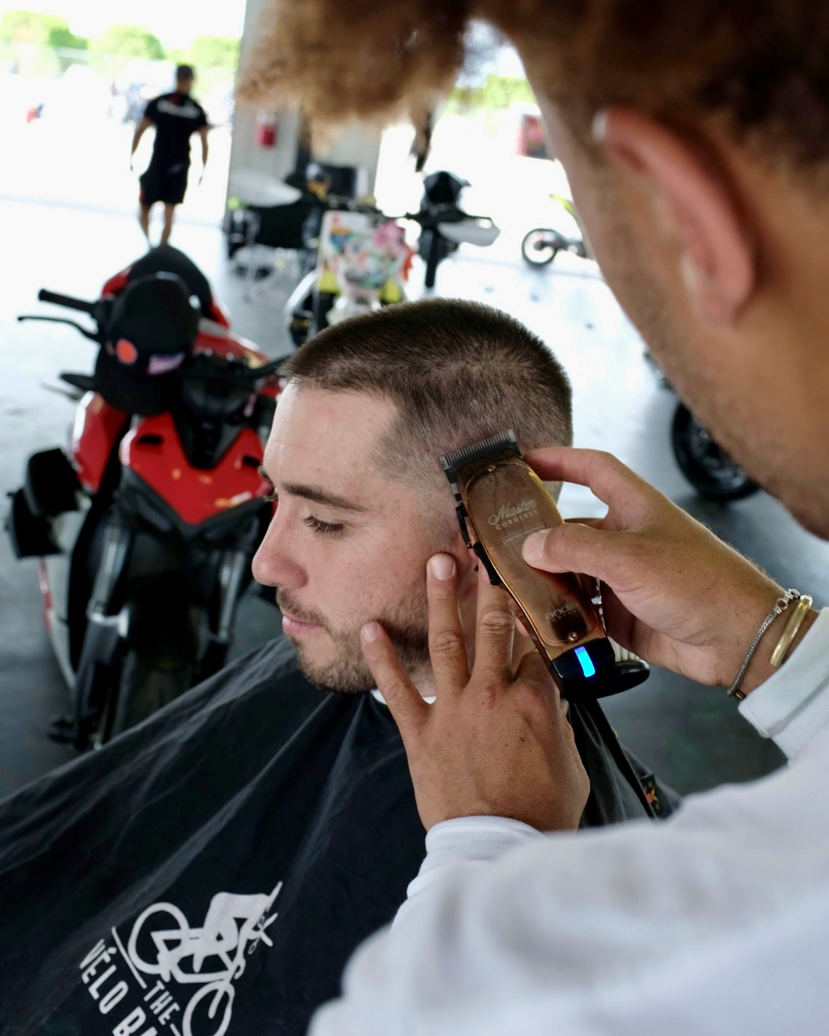 Side of Julien Howard's face and hands holding Andis Master Clipper in the foreground and using them on customer with a motorcycle in the background